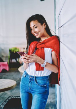 İçerik sahibi, uzun siyah saçlı, gündelik giysiler içinde, yaratıcı çalışma alanındaki ahşap duvara yaslanmış cep telefonlarını izleyen mutlu bir etnik kadın.
