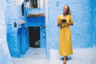 Sarı elbiseli, gülümseyen sarışın kadın elinde fotoğraf makinesiyle antik bir caddede Fas 'ta mavi duvarları olan eski taş binanın yanında duruyor.