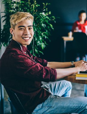 İyimser gülümseyen, kulaklıklı, kulaklık takan, iş yerindeki masada oturup kameraya bakarak dizüstü bilgisayar kullanan Asyalı adamın yan görüntüsü.