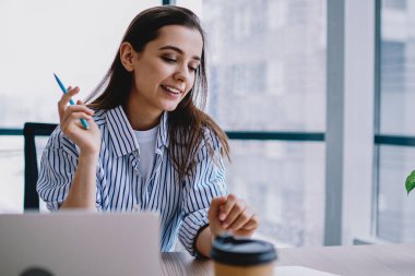 Modern ofisin penceresinde masa başında otururken günlük kıyafetlerle gülümseyen ve açık planlamacıda notlar okuyan mutlu genç kadın.