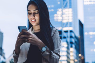 Şehir caddesinde gökdelenlerin önünde duran, resmi kıyafetler içinde, ağzı açık akıllı telefon kullanan çekici, modern bir kadın.