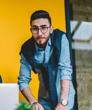 Modern bilgisayar ile masada oturan ofis yönetimi hakkında yönetici ile fikir tartışırken kamera bakarak vizyon düzeltme için optik gözlük ciddi erkek kurumsal işçi portresi