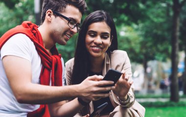 Gözlüklü, şık giyinmiş, akıllı telefondan sevgiliye gösteren bir erkek arkadaş. İkisi de arka planda bulanık bir şekilde parkta oturuyor.