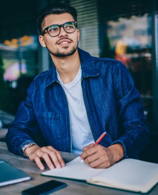 Gözlük pensive sakallı adam kafe iç dizüstü bilgisayar ile oturan blog da yayın için not defteri fikirleri yazma, gözlük uzakta notlar bakarak gülümseyen erkek öğrenci hayal