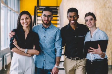 Açık modern ofis binasında renkli koridorun iç kısımları bulanık dururken kameraya gülümseyen ve kucaklaşan resmi kıyafetli neşeli yetişkin çalışanlar.