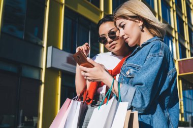 Güneş gözlüklü, alışveriş torbalarıyla gezen şık bir kadın ve şehir merkezinin dışındaki akıllı telefonlara bakıyor.