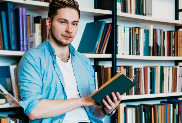 Confident charismatic young man in casual wear choosing book for homely reading sitting on chair about bookshelf in library looking at camera