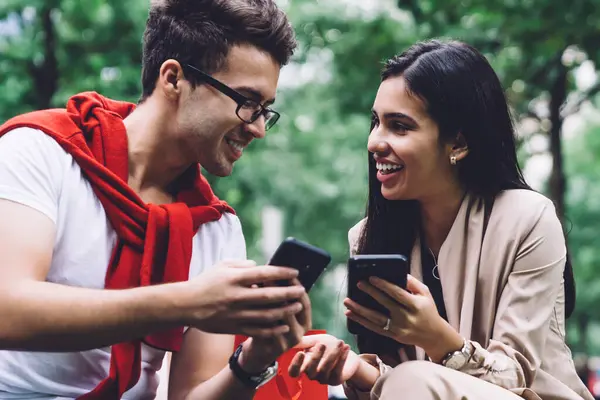 Cheerful and carefree couple in casual clothes sitting on stone bench in quiet park friendly talking and showing screens of smartphones