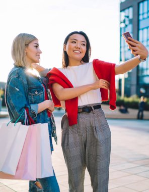 Alışveriş torbalarıyla çok kültürlü ve çekici genç bayan arkadaşları akıllı telefonlarıyla alışveriş merkezinin yakınında dururken selfie çekiyorlar.