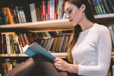 Uzun siyah saçlı, entellektüel, kısa etekli ve beyaz kazaklı bir kadın kütüphanede yerde oturmuş kitap okuyor.