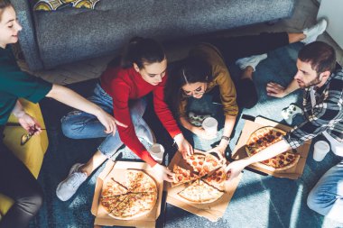 Yukarıdan gülümseyen mutlu insanlar gündelik kıyafetlerini giyip pizzalarını paylaşırken güneşli, modern bir odada kanepede dinleniyorlar.