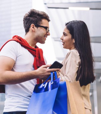 Akıllı telefon kullanırken, alışveriş torbalarıyla şehir binasının yanında dinlenirken ve arka planda bulanık bir şekilde birbirine bakarken çok ırklı çiftleri sevmenin düşük açılı yan görüntüsü.