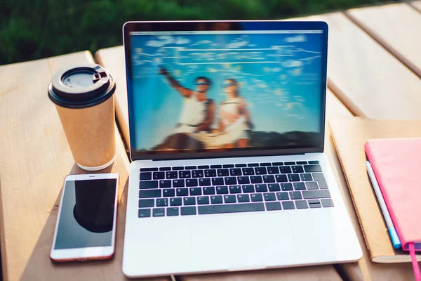 Open laptop computer with blurred colourful wallpaper on screen standing outdoors on wooden table with take away coffee cup, notebook and smartphone. Freelancer workplace on fresh air. Dream desktop