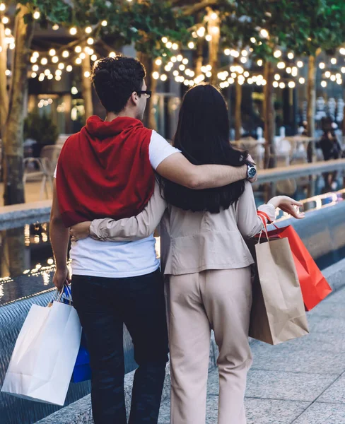 Back view of modern trendy couple in casual clothes hugging with love while walking on city street with shopping bags