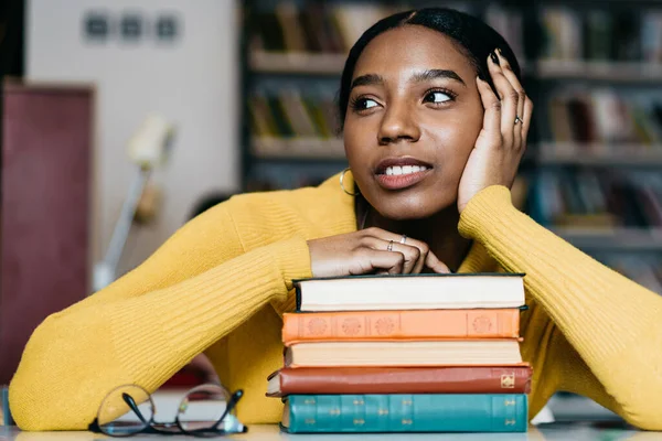 Pensive African American student in casual clothes looking away and contemplating while sitting at table with books and leaning on hand against blurred bookshelf in library