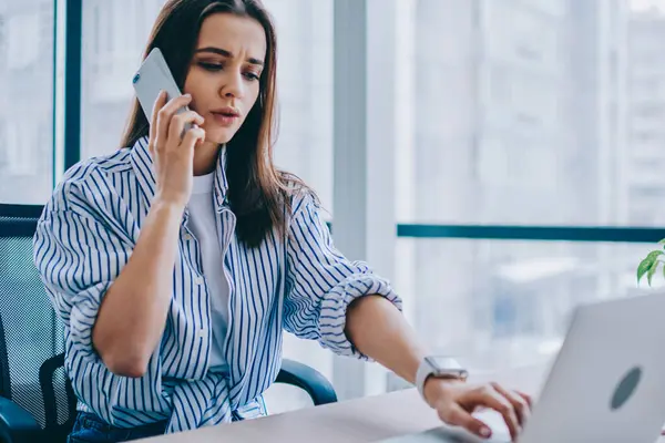 Focused young female in casual outfit answering phone call and browsing laptop while sitting at table and working against window in modern office