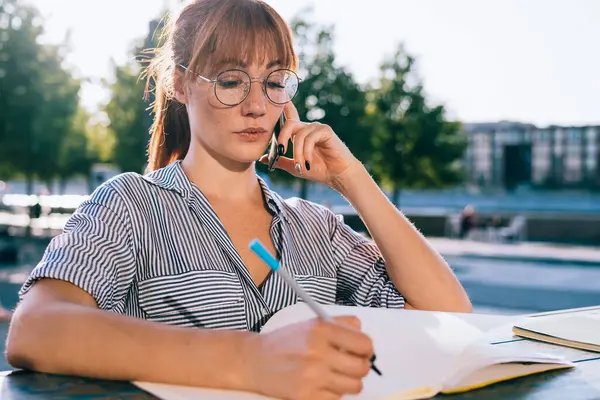 Concentrated young female student in eyeglasses and casual shirt writing in notebook while preparing for university exam in city park in sunny summer day