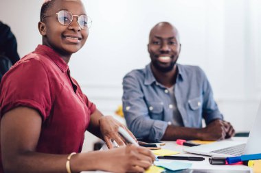 Gülümseyen siyahi meslektaşlar dizüstü bilgisayarlarıyla masada otururken erkekler internette sörf yapıyor ve kadınlar not alıyor ve bulanık arka plandaki kameraya bakıyor.
