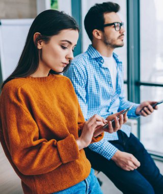 Cep telefonuyla mesajlaşan çekici genç bir bayan ve gözlüklü bir erkek arkadaş akıllı telefon kullanarak hafif ofisteki masada yan yana yatıyorlar.