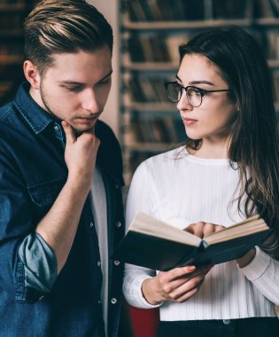 Kitaplığın karşısındaki kütüphanede kot gömlekli bir adamla duran beyaz kazaklı genç kadın kitabı işaret ediyor.