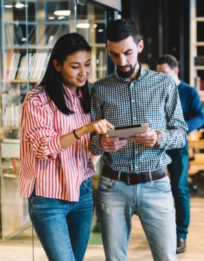 Zeki çoklu etnik kadın ve erkek, ahşap masalar ve öğrencilerle dolu geniş bir kütüphanede kağıt tablet çizmekten bahsediyorlar.
