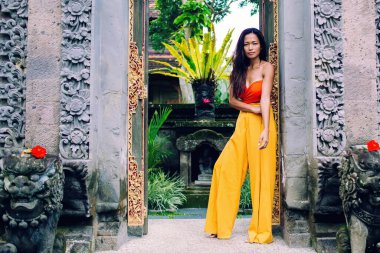 Kırmızı tişörtlü ve sarı pantolonlu Asyalı genç bayan antik egzotik tapınağın kemerinin önünde duruyor.