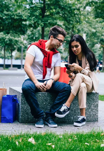 Sweet couple chilling on stone bench near paper bags after shopping and using smartphone outside in park on blurred background