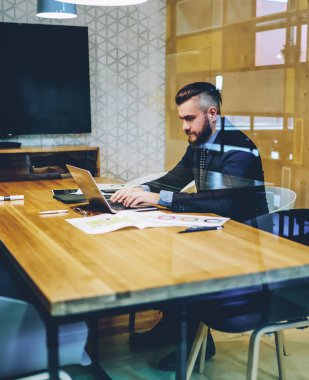 Resmi giyinmiş dalgın işadamı, cam duvarın arkasında kendi ofisinde oturan modern laptop cihazında klavye bilgisi takıyor. Web sitesindeki arama bilgileri için bilgisayarda çalışan erkek tüccar