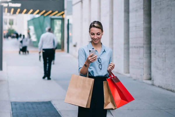 Happy female shopper with paper bags in hand enjoying recreation after buying purchases making online booking for taxi cab, cheerful hipster girl chatting with friend texting message about sales