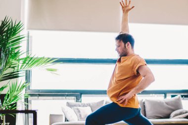 Spor yapan genç bir adamın sabah evde çalışırken el ele tutuşması. Hippi adam yoga eğitiminden önce ısınıyor. Havalı halı ve ahenk ve sağlıklı zindeliğin keyfini çıkarıyor.