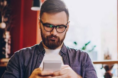 Görme düzeltmesi için gözlük takan ciddi bir erkek blogcu akıllı telefon aygıtında SMS 'leri okurken aynı zamanda laptoplu bilgisayarla birlikte kafenin iç kısmında vakit geçiriyor.