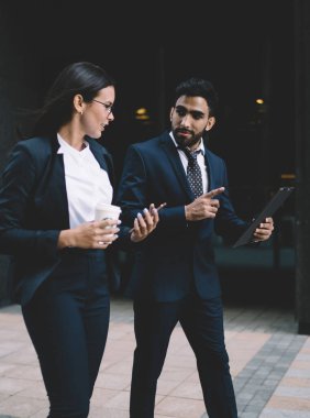 Resmi takım elbise giymiş iki farklı olgun ekonomist arka planda modern heykellerle finans bölgesinde dolaşıyor ve ofis binasına giderken klasörden muhasebe raporlarını tartışıyorlar.