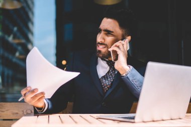 Bilmece deneyimli girişimci resmi olarak muhasebe raporlarındaki bilgi verilerini tartışırken akıllı telefon aygıtıyla bilgisayarın başında bilgisayarla otururken iş ortağıyla konuşuyor.