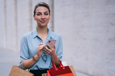 Şehir ortamında ayakta duran ve internet sitesinde fiyatları, teknoloji ve iletişim kavramını kontrol etmek için akıllı telefon cihazıyla kamusal kablosuz bağlantı yakalayan mutlu kadın alışverişi portresi