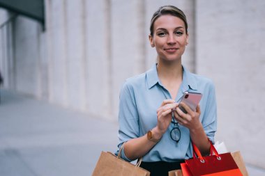 Şehir ortamında ayakta duran ve internet sitesinde fiyatları, teknoloji ve iletişim kavramını kontrol etmek için akıllı telefon cihazıyla kamusal kablosuz bağlantı yakalayan mutlu kadın alışverişi portresi
