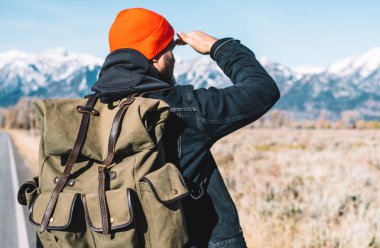 Yürüyüş için şık sırt çantasıyla asfalt yolda dikilirken iyi bir ritm ve tempoyla tepeye çıkmak için dağlara giden yolu arayan genç erkek yürüyüşçünün arka görüntüsü, nefes kesici kavram.