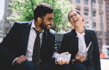 Heyecanlı erkek ve kadın iş arkadaşları finans bölgesinde arkadaşlığın tadını çıkarıyorlar açık havada oturup güneşli bir günde şakalar yaparak gülüyorlar, iş adamları kavramı ve mutluluk.