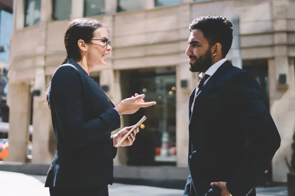 Successful smiling woman boss talking to male spanish employee about schedule for day during break in business district, positive colleagues in elegant wear enjoying live communication outdoors
