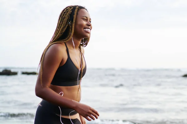 Cheerful african female runner dressed in sport top listening favourite music songs in modern earphones during sport workout walking on sea coastline of tropical island, concept of healthy lifestyle