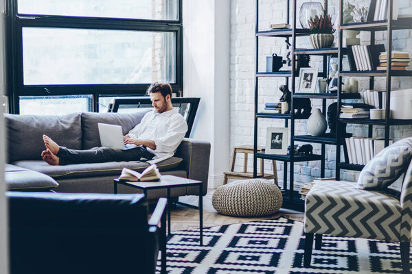 Skilled male freelancer taking rest on comfortable couch while working remotely in stylish home interior, young hipster guy writing email while lying on sofa with modern laptop computer indoors