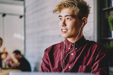 Başarılı Asyalı erkek serbest çalışan, modern dizüstü bilgisayar, eğlence ve keyif kavramı ile uzaktan çalışırken elektronik ekipmanlarla favori müzik listesini dinlerken başka yöne bakıyor.