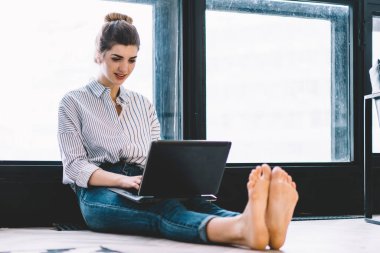 Gülümseyen genç bir kadın dizüstü bilgisayarda klavye kullanıyor ve uzaktan kumanda uygulamasını güncelliyor. Büyük pencereli, modern bir dairede yerde oturuyor. Hipster kızı netbook üzerine ders için metin yazıyor.