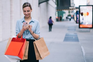 Neşeli beyaz kadın blog yazarı cep telefonuyla 4G internet bağlantısı kullanarak sosyal ağda yayın yapıyor. Alışveriş paketleriyle arkadaşlarıyla akıllı telefon aracılığıyla mesajlaşan mutlu hippi kız.