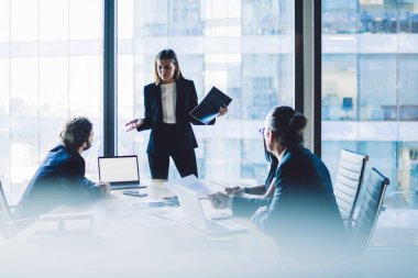 Şaşırmış kadın CEO konferans odasında çalışanlarla birlikte sunum taşıyor. Toplantı masasının yanında duran kadın yönetici, gökdelenin içindeki toplantı sırasında stratejiyi açıklarken,