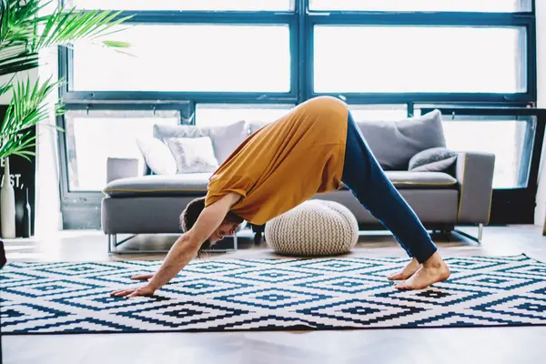 Motivated young man standing in yoga pose on carpet in modern apartment with stylish interior.Hipster guy in sportive wear leads healthy lifestyle during morning sportive exercises at home
