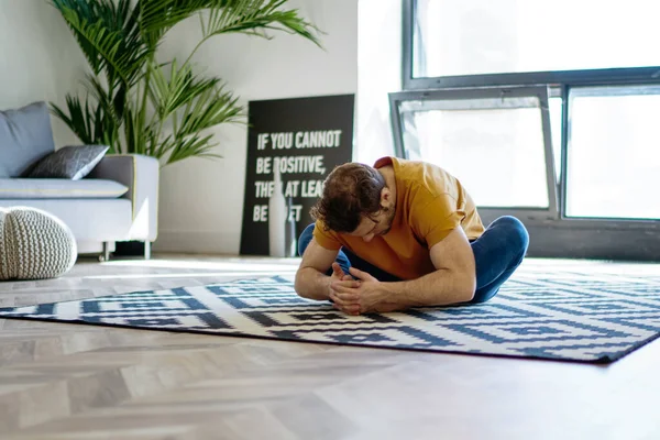 Healthy young man sitting in pose lotus on carpet and doing yoga exercises during morning workout in modern apartment.Hipster guy practicing meditation in stylish flat with home interior