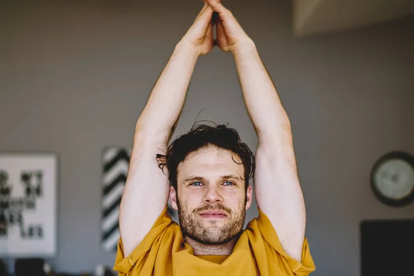 Handsome young man with blue eyes holding hands in namaste above the head above the head during meditation and relaxation in modern apartment.Motivated hipster guy doing yoga exercises during training