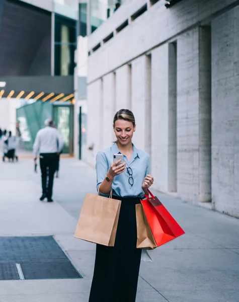 Happy female shopper with paper bags in hand enjoying recreation after buying purchases making online booking for taxi cab, cheerful hipster girl chatting with friend texting message about sales
