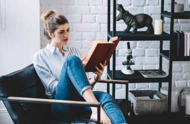 Düşünceli genç bir kadın sandalyede oturup edebiyat kitabı okuyor ve modern bir dairede keyfine bakıyor. Gündelik giyinmiş hippi kız, elinde kitapla evde şık bir iç mekan ile rahatlıyor.