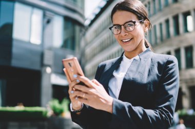 Zarif bir takım elbise giymiş, şık gözlüklü, gülümseyen, gözlüklü, neşeli kadın avukatın yarım boy portresi finans bölgesinde güneşli bir günün tadını çıkarıyor.